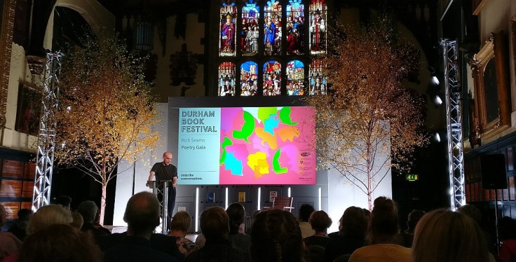 Andrew McMillan introducing the poets at Durham Book Festival, on a stage surrounded by trees with a stained glass window behind